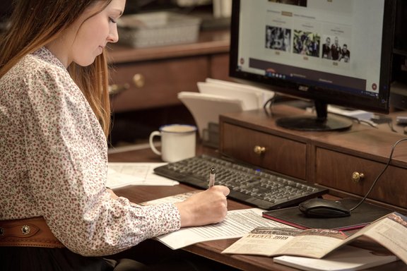 Frau sitzt an einem Schreibtisch mit Computer und schreibt auf ein Blatt