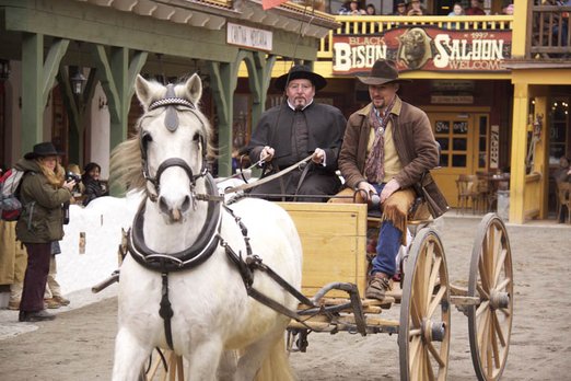 Pfarrer und Cowboy sitzen auf einer Kutsche, die von einem Pferd durch die Main Street der Westernstadt gezogen wird