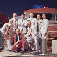 Acht Männer in Anzügen vor einem alten Fahrzeug