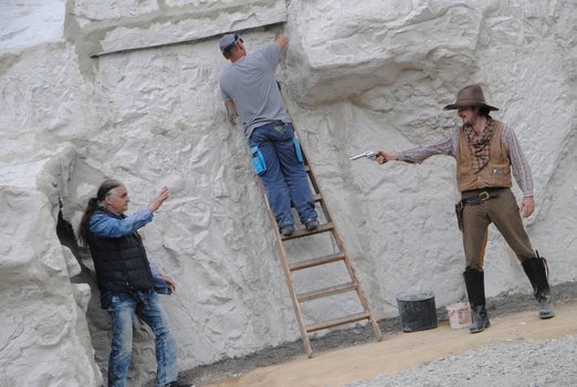 Mann steht auf einer Leiter am Fels, während ein Cowboy mit einem Revolver auf einen Indianer zielt