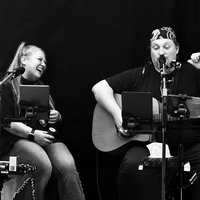 Ein Musiker mit einer Gitarre und eine Sängerin sitzen auf der Bühne, umgeben von Nebel und Lichtern, während sie ein Lied performen.