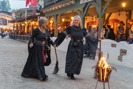Zwei Frauen in Hexenkostümen mit Besen gehen auf ein kleines Feuer zu