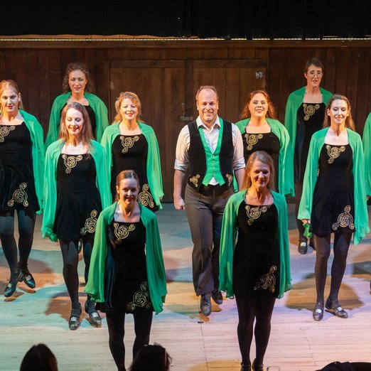 Tanzgruppe in schwarzen Kleidern und grünem Umhang auf der Bühne
