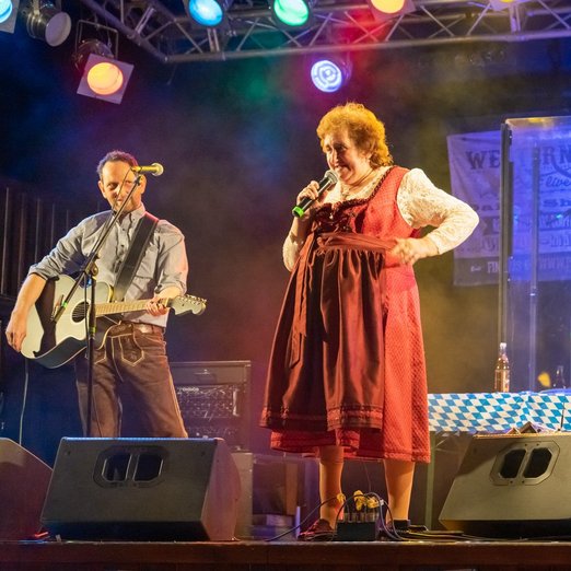 Frau in rotem Dirndl und Mann in Lederhose mit Gitarre auf einer Bühne
